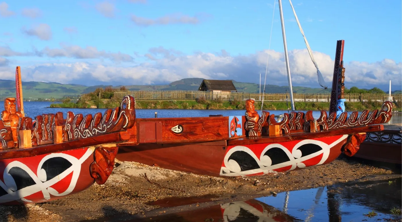 Two carved waka taua war canoes drawn up on the shore of Lake Rotorua under a blue sky