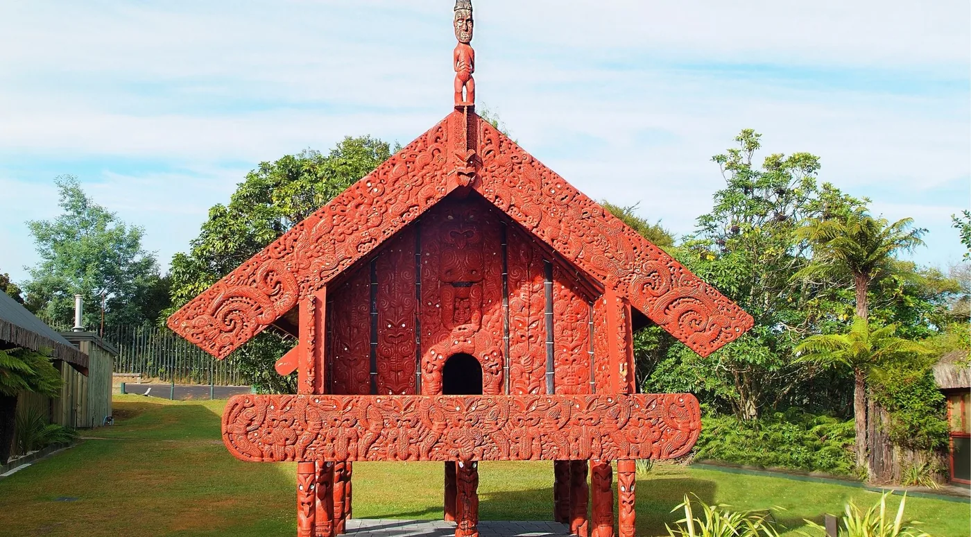An ornately carved red pātaka storehouse raised on carved posts against a blue sky