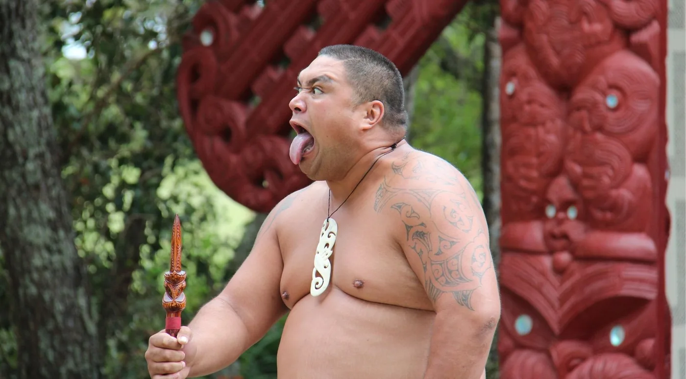 A performer with a taiaha issuing a wero challenge in front of a red carved meeting house