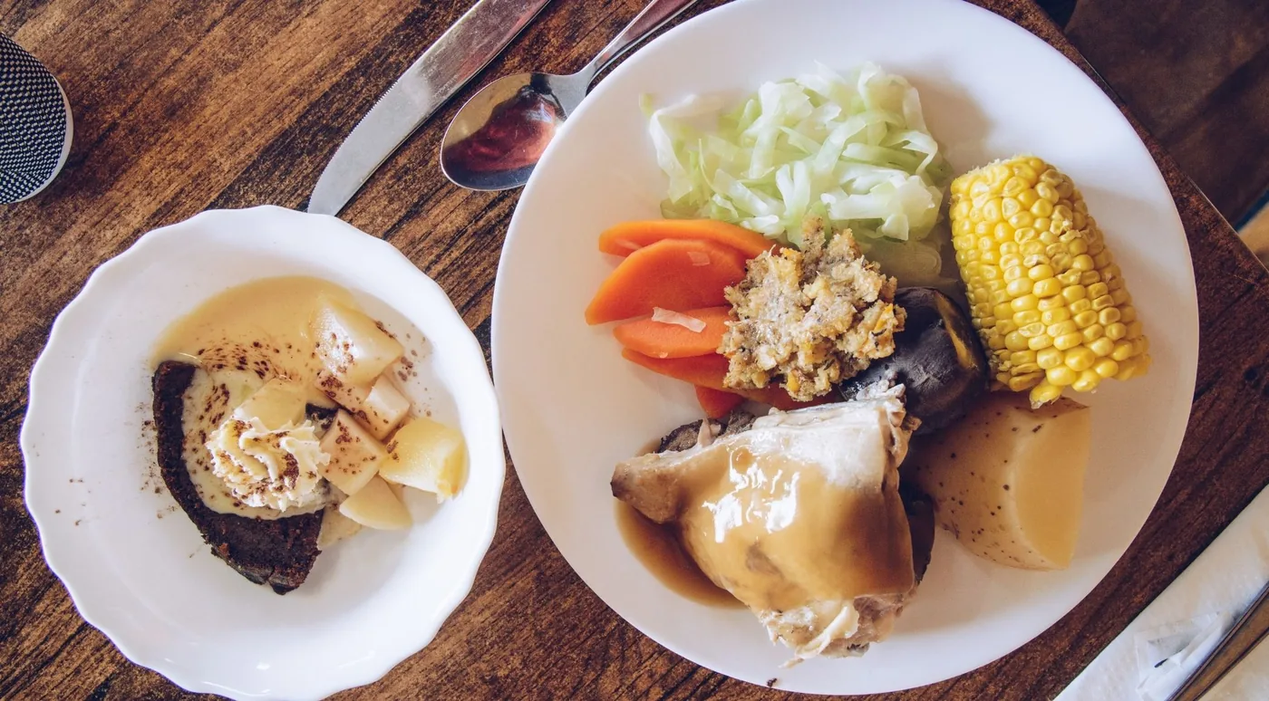 A plated hāngī meal of chicken, kūmara, potato, cabbage, corn and stuffing beside a steamed pudding